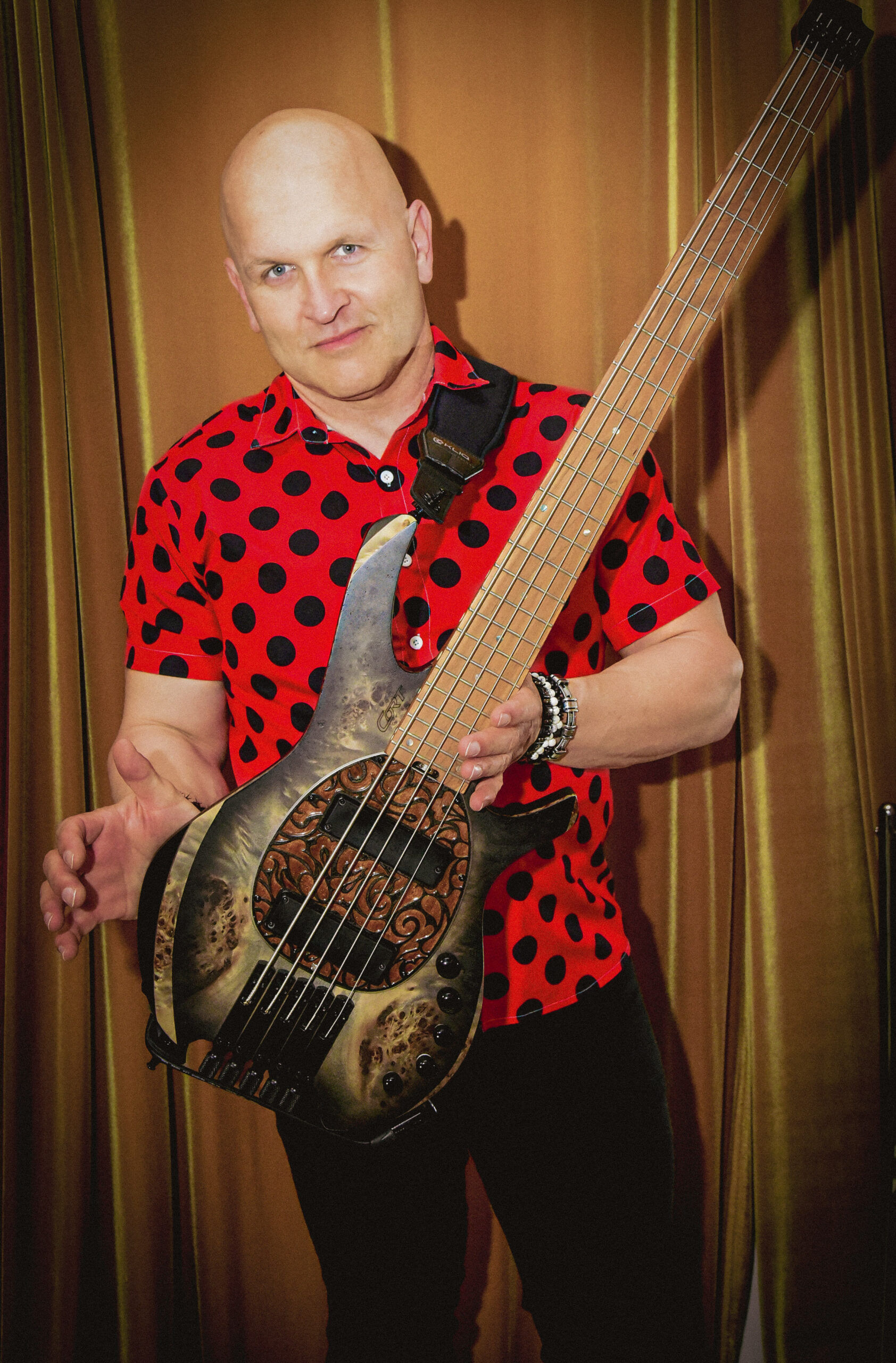 Danny Knapp in a red polka-dot shirt holding a uniquely designed bass in front of gold curtains.