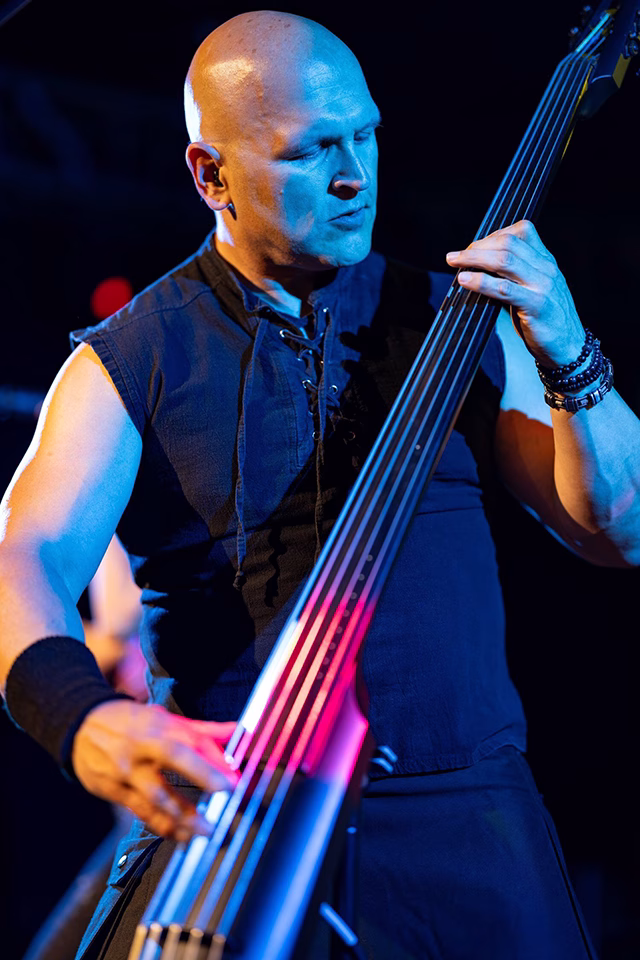 Danny Knapp in a sleeveless black shirt playing an upright electric bass on stage under blue and pink lighting.